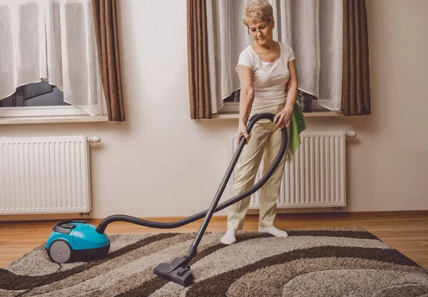 Una anciana haciendo tareas de mujeres en casa. Vacumming la alfombra —  Fotos de Stock