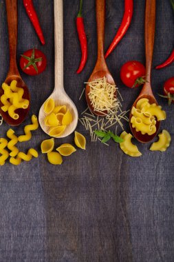 Kaşık koyu ahşap arka plan, üstten görünüm üzerinde farklı makarna. Pişmemiş makarna, biber, domates ve fesleğen yakın çekim.