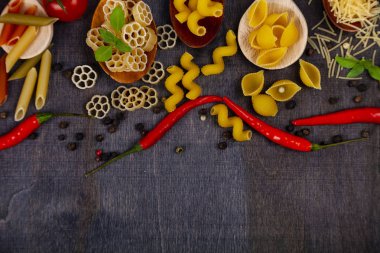 Kaşık koyu ahşap arka plan, üstten görünüm üzerinde farklı makarna. Pişmemiş makarna, biber, domates ve fesleğen yakın çekim.