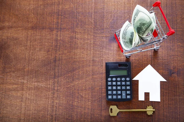 Money in a shop trolley,calculator, key and a house on a wooden background.. Buying a property.