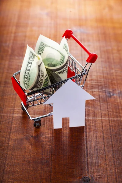 Money in a shop trolley and a house on a wooden background.. Buying a property.