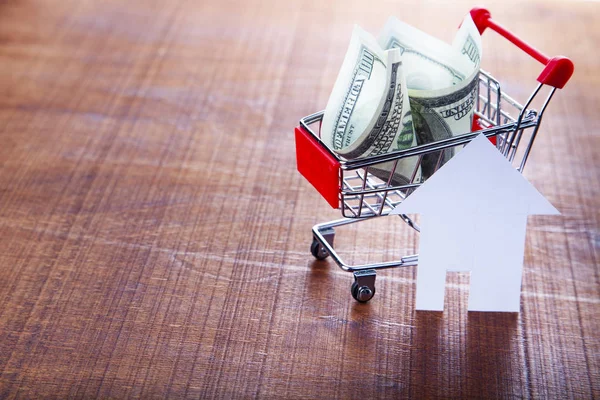 Money in a shop trolley and a house on a wooden background.. Buying a property.