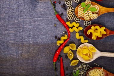 Kaşık koyu ahşap arka plan, üstten görünüm üzerinde farklı makarna. Pişmemiş makarna, biber, domates ve fesleğen yakın çekim.