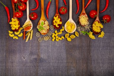Kaşık koyu ahşap arka plan, üstten görünüm üzerinde farklı makarna. Pişmemiş makarna, biber, domates ve fesleğen yakın çekim.