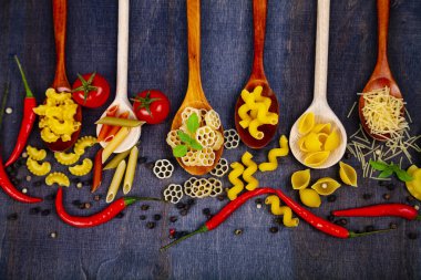 Kaşık koyu ahşap arka plan, üstten görünüm üzerinde farklı makarna. Pişmemiş makarna, biber, domates ve fesleğen yakın çekim.