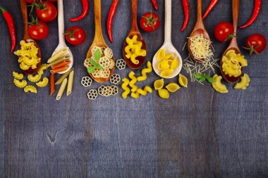 Kaşık koyu ahşap arka plan, üstten görünüm üzerinde farklı makarna. Pişmemiş makarna, biber, domates ve fesleğen yakın çekim.
