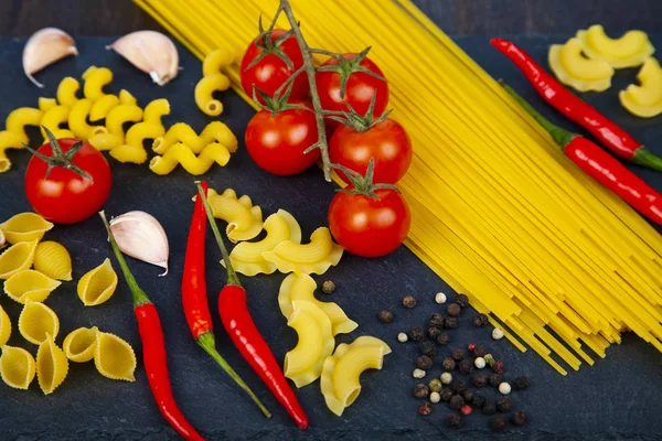 Different pasta on a wooden background. Raw pasta, chili, tomatoes and basil close-up.