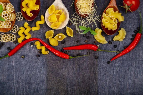 Kaşık koyu ahşap arka plan, üstten görünüm üzerinde farklı makarna. Pişmemiş makarna, biber, domates ve fesleğen yakın çekim.