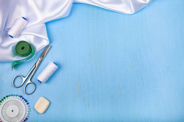 Mavi ahşap arka plan üzerinde dikiş aksesuarları. Beyaz saten kumaş, makas ve iş parçacığı.