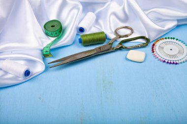 Mavi ahşap arka plan üzerinde dikiş aksesuarları. Beyaz saten kumaş, makas ve iş parçacığı.