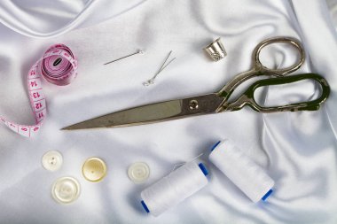 Beyaz saten kumaş üzerine dikiş aksesuarları. Beyaz saten kumaş, makas, iplik, yüksük, iğne ve ölçüm bandı.