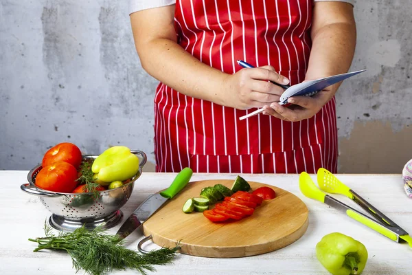 Woman in a red apron writes in a notebook in the kitchen. Recipe for eating