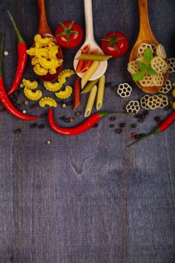 Kaşık koyu ahşap arka plan, üstten görünüm üzerinde farklı makarna. Pişmemiş makarna, biber, domates ve fesleğen yakın çekim.