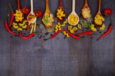 Kaşık koyu ahşap arka plan, üstten görünüm üzerinde farklı makarna. Pişmemiş makarna, biber, domates ve fesleğen yakın çekim.