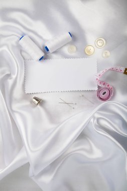 Dikiş aksesuarları ve beyaz saten kumaş üzerine bir not. Beyaz saten kumaş, iplik, yüksük, iğne ve ölçüm bandı.