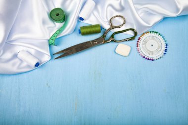 Mavi ahşap arka plan üzerinde dikiş aksesuarları. Beyaz saten kumaş, makas ve iş parçacığı. Metniniz için yer.