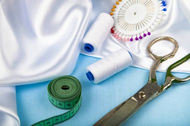 Mavi ahşap arka plan üzerinde dikiş aksesuarları. Beyaz saten kumaş, makas ve iş parçacığı.