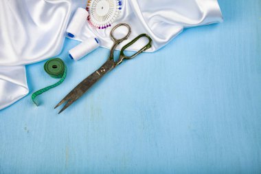Mavi ahşap arka plan üzerinde dikiş aksesuarları. Beyaz saten kumaş, makas ve iş parçacığı. Metniniz için yer.