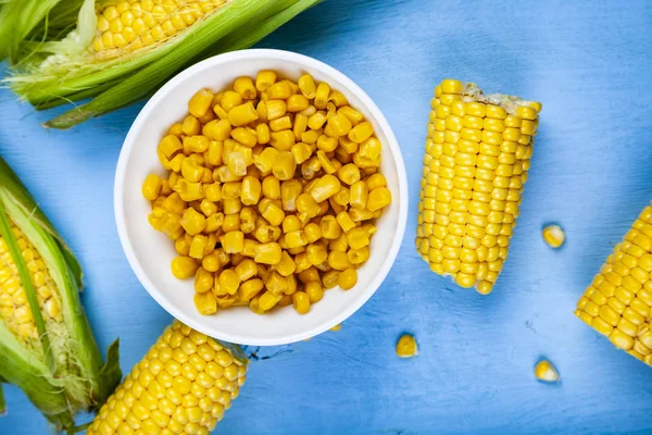 Ripe corn on a blue table close-up. Raw and canned corn in a white bowl, top view.