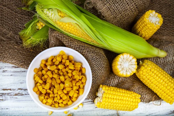 Ripe yellow corn and canned corn in a white bowl on a wooden table. Yellow ripe corn, top view