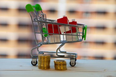 Alışveriş arabasında bir ev ve inşaat halindeki bir binanın arka planında para. Bir mülk satın almak.