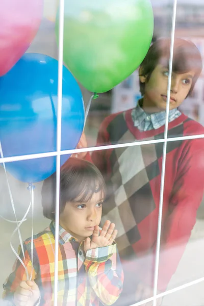 Dos hermanos felices sosteniendo globos detrás del cristal de la ventana —  Fotos de Stock