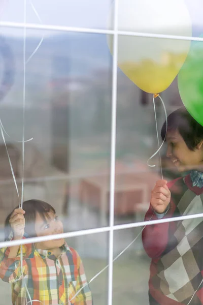 Dos hermanos felices sosteniendo globos detrás del cristal de la ventana —  Fotos de Stock