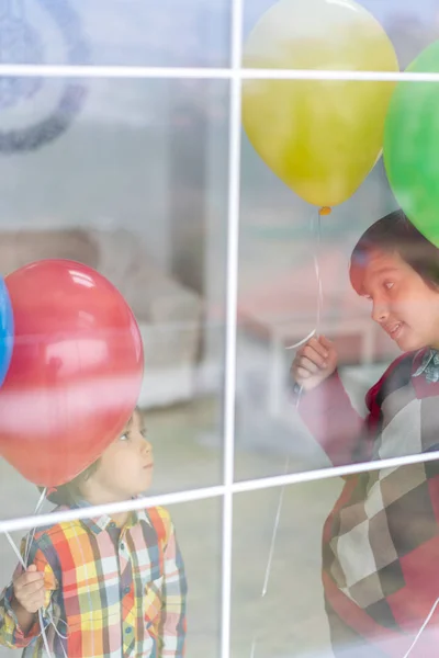 Dos hermanos felices sosteniendo globos detrás del cristal de la ventana — Foto de Stock