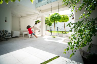 Küçük bir çocuk bir villanın arka bahçesinde salıncakta oynuyor.