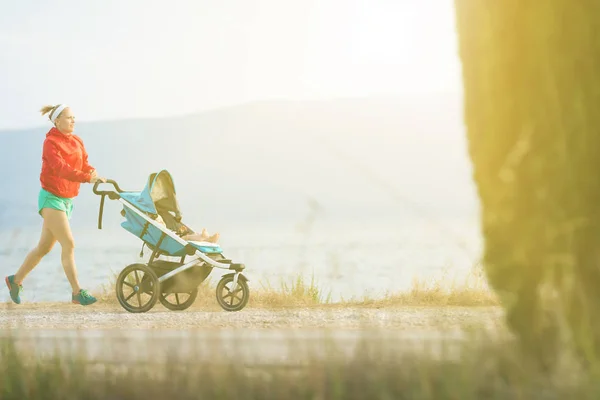 Running super mother with child in baby stroller enjoying sunset and mountains landscape. Jogging or power walking supermom, active family at sunset. Beautiful inspirational mountains landscape.