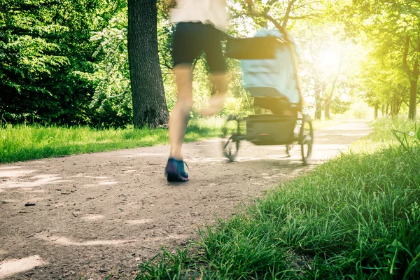 Running woman with baby stroller enjoying summer in park. Jogging or power walking supermom, active family with baby jogger, motion blur.