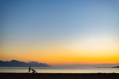 Anne Bebek arabası annelik günbatımı manzara, zevk ile silüeti. Kadın çocuk arabası ile gün batımında sahilde koşu veya yürüyüş. Güzel ilham verici Dağları manzara.