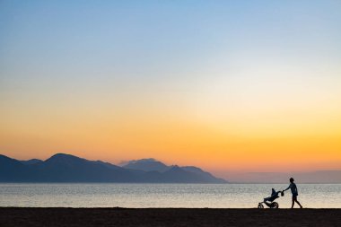 Anne Bebek arabası annelik günbatımı manzara, zevk ile silüeti. Kadın çocuk arabası ile gün batımında sahilde koşu veya yürüyüş. Güzel ilham verici Dağları manzara.