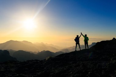 Couple hikers celebrating success in sunset mountains, accomplish with arms up outstretched. Young man and woman looking at beautiful inspirational landscape view, Gran Canaria Canary Islands. clipart