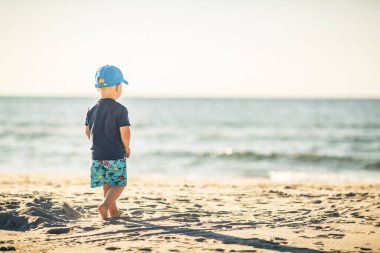 Güneşli bir sahilde yürüyen küçük bir çocuk. Küçük çocuk kumda yürüyor. Güzel ilham verici sahil ve okyanus manzarası, manzara.