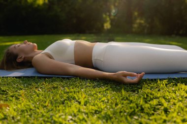 Spor giyim bölümünde genç bir kadın parktaki spor minderinde vücut esnetiyor, yoga yapıyor. Savasana. Sağlıklı yaşam tarzı, spor. Fitness. Sabah parkında yoga yapan kız. Dizinin bir parçası. Yumuşak odak.
