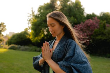 Kadın halka açık bir parkta yoga ve meditasyon yapıyor, gözleri kapalı rahatlıyor. Dikkatli meditasyon konsepti. Sağlıklı olmak. Odak bulanıklığı ve gürültü etkisi yok. Dizinin bir parçası.