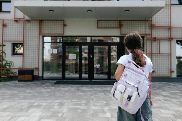 Задний вид на мультиэтнических школьниц младших классов ген Альфа одноклассников с рюкзаком в повседневной одежде обнимают друг друга стоя на улице. Часть серии.