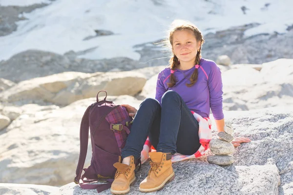 肖像快乐的女孩徒步旅行者与背包坐在岩石上的雪和山在背景 — 图库照片