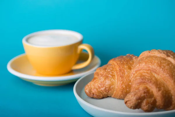 Gelbe Tasse mit Croissant auf blauem Hintergrund — Stockfoto