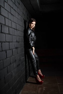 Deri elbiseli genç kız karanlık duvarın önünde duruyor.