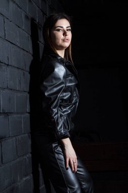 Deri elbiseli genç kız karanlık duvarın önünde duruyor.