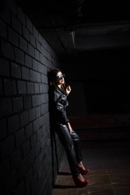 Deri elbiseli genç kız karanlık duvarın önünde duruyor.