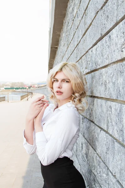 年轻的金发姑娘穿着白衬衫站在花岗岩墙边 — 图库照片