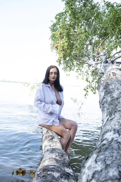 young girl in a white wet shirt stands on a birch tree fallen into the water