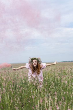Çelenk takmış bir kız ve renkli dumanla tarlada dans ediyor.