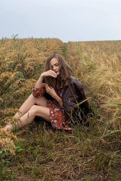 ドレスを着た若い女の子と革のジャケットが大麦の熟した耳の間のフィールドに座っている — ストック写真