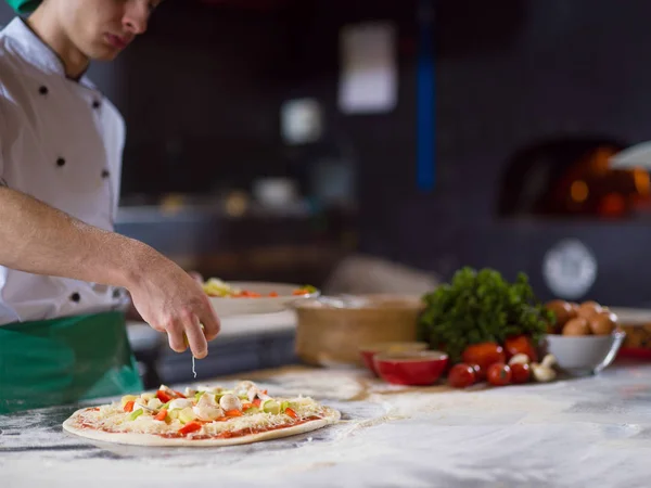 chef putting fresh vegetables over pizza dough on kitchen table