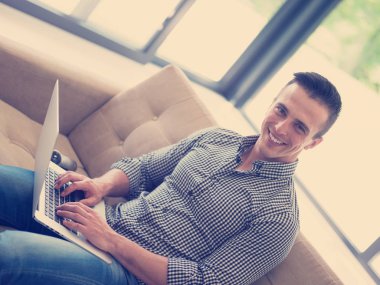 Genç, yakışıklı adam koltukta dizüstü bilgisayar kullanıyor.