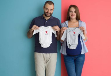 Beautiful pregnant woman and her husband expecting baby holding baby bodysuits and smiling over colorful background clipart